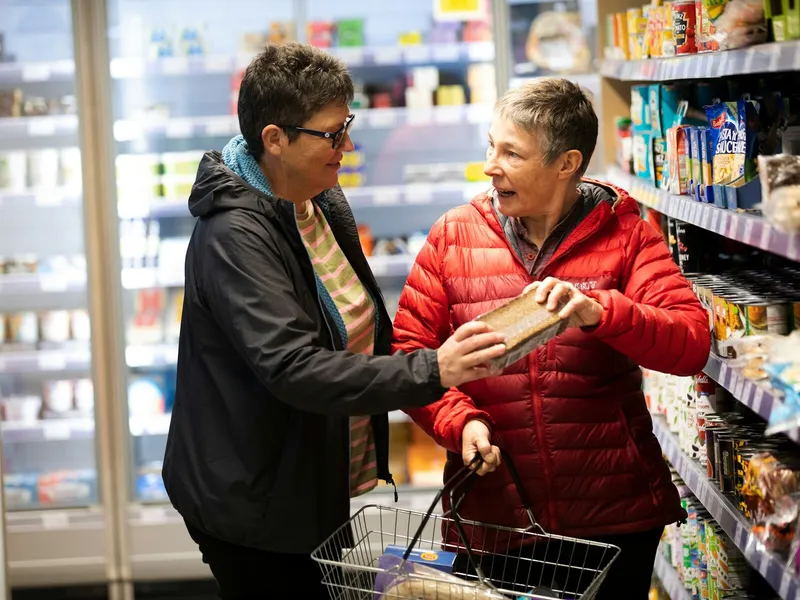 Two women choose groceries together in a shop, holding a loaf beside their shopping basket.
