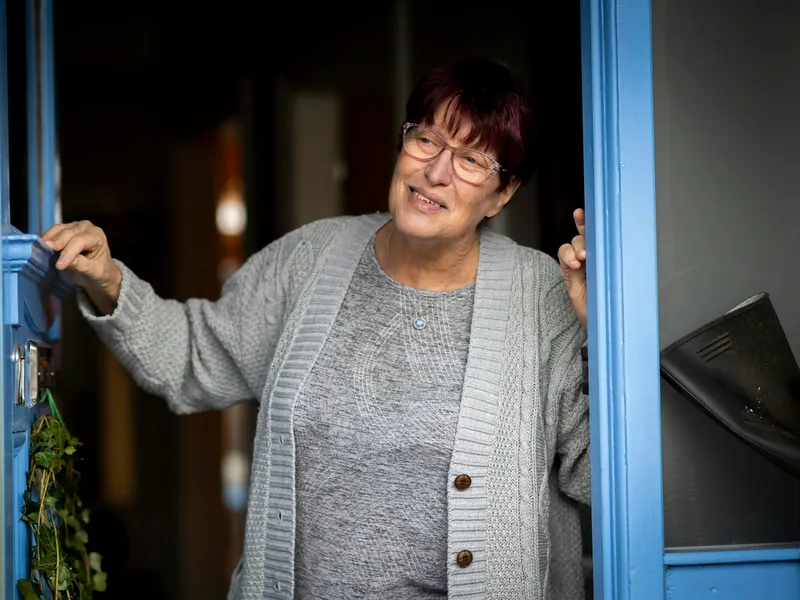 A woman opens her blue front door and smiles at the person arriving.